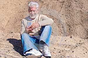 Attractive senior man  sitting on the clear rocks enjoying the sun in a winter day - using mobile phone and smiling. Relaxed