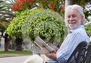 Attractive senior man with white beard smiles sitting in the park reading a book surrounded by flowering plants and palms -