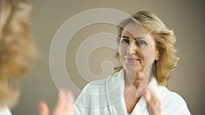 Attractive senior woman brushing hair in front mirror and smiling, aged beauty