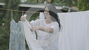 Attractive senior woman with long hair black hair hanging white clothes on a clothesline outdoors. Washday. Positive