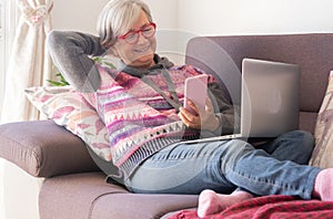 Attractive senior woman resting on sofa at home using laptop and mobile phone. Smiling elderly retiree enjoying tech and social