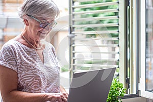 Attractive senior woman sitting close to the window using laptop computer. Smiling caucasian female enjoying technology and social
