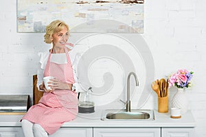 attractive senior woman sitting on kitchen counter with cup of coffee