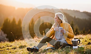 Attractive senior woman sitting outdoors in nature at sunset, relaxing with coffee.