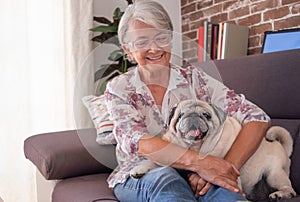 Attractive senior woman sitting on sofa at home embracing her old pug dog lying on her legs