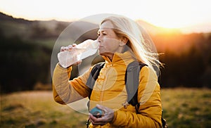 Attractive senior woman walking outdoors in nature at sunset, drinking water.