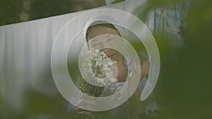 Attractive senior woman with white shawl on her head tears off daisy petals at the clothesline outdoors. Washday