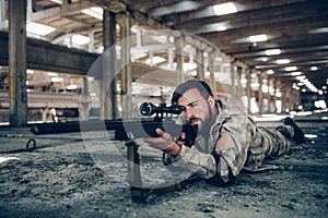 Attractive and serious guy is lying on the ground in a big hangar. He is taking aim. Man in looking through lens. He is
