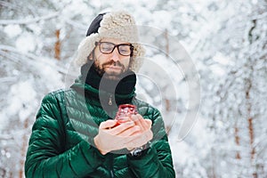 Attractive serious male wears warm winter clothes, keeps candle, stands against winter forest background, enjoys fresh air, being