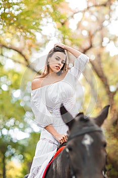 Horseback riding. Beautiful young woman in a white dress riding on a brown horse outdoors.