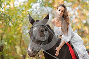 Horseback riding. Beautiful young woman in a white dress riding on a brown horse outdoors.