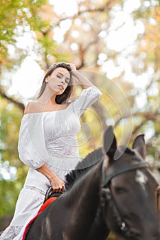 Horseback riding. Beautiful young woman in a white dress riding on a brown horse outdoors.