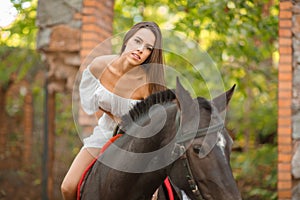 Horseback riding. Beautiful young woman in a white dress riding on a brown horse outdoors.