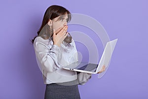 Attractive shocked businesswoman with laptop in hands posing isolated over lilac background, wearing elegant attire, has