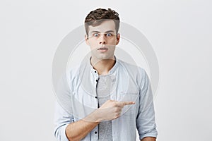 Attractive shocked young caucasian male with bugged eyes dressed casually pointing index finger sideways at grey blank