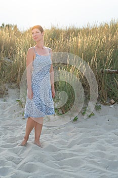 Attractive short haired woman in her thirties, dressed in light summer dress standing barefoot on the beach with grass