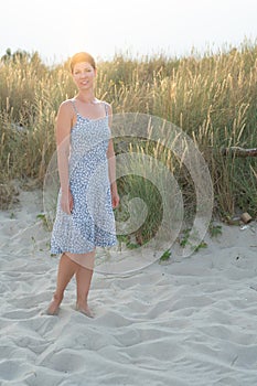 Attractive short haired woman in her thirties, dressed in light summer dress standing barefoot on the beach with grass