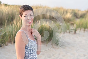 Attractive short haired woman in her thirties, dressed in light summer dress standing barefoot on the beach with grass