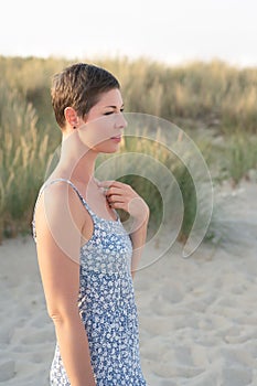 Attractive short haired woman in her thirties, dressed in light summer dress standing barefoot on the beach with grass