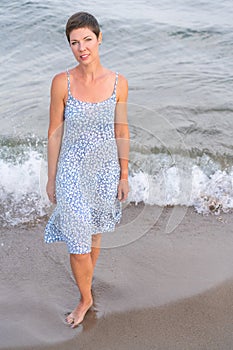 Attractive short haired woman in her thirties wear in light summer dress walks barefoot on the beach with sea in the