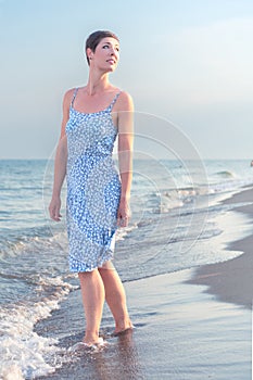 Attractive short haired woman in her thirties wear in light summer dress walks barefoot on the beach with sea in the