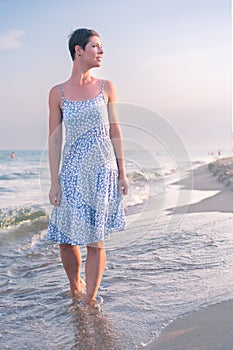 Attractive short haired woman in her thirties wear in light summer dress walks barefoot on the beach with sea in the