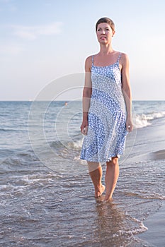 Attractive short haired woman in her thirties wear in light summer dress walks barefoot on the beach with sea in the