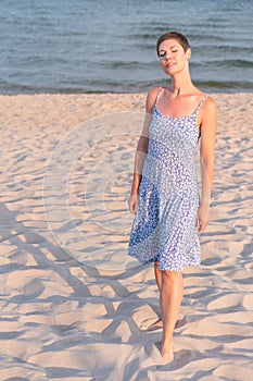 Attractive short haired woman in her thirties wear in light summer dress walks barefoot on the beach with sea in the