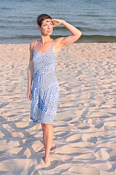 Attractive short haired woman in her thirties wear in light summer dress walks barefoot on the beach with sea in the