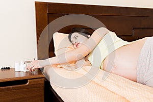 Attractive sick pregnant woman is lying in sofa. Taking pills from colds