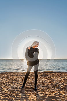 Attractive slender person girl blonde stands posing on the shore of the water in black clothes with a background of the rising sun
