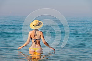 An attractive slender young Caucasian woman in a swimsuit and a straw hat stands waist-deep in sea water in the morning
