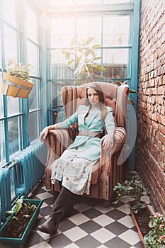 Attractive slim blonde woman in blue jacket, floral dress and cowboy boots sits in a vintage armchair in a loft interior