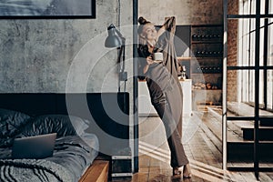 Attractive slim caucasian young woman in satin pajama with cup of coffee in her hand stretching in bedroom