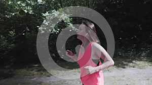 Attractive slim girl in bright pink top with headphones is breathing heavily during training. Young sportive woman running in park