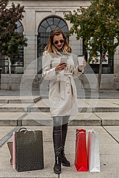 Attractive slim woman looks at her mobile phone while having a coffee in the street next to her shopping bags