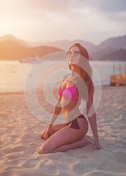 Attractive slim young brunette woman in bikini and sunglasses sitting on beach smiling admiring sunrise with mountains