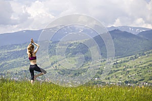 Attractive slim young woman doing yoga exercises outdoors on background of green mountains on sunny summer day