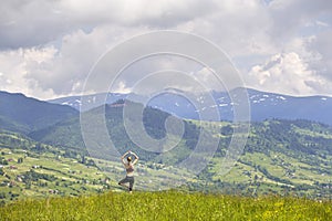 Attractive slim young woman doing yoga exercises outdoors on background of green mountains on sunny summer day