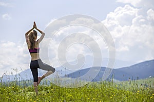 Attractive slim young woman doing yoga exercises outdoors on background of green mountains on sunny summer day