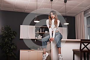 Attractive slim young woman model in a fashion white top in vintage jeans in trendy sneakers sexually sits on the kitchen table in