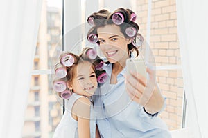 Attractive small kid with positive expression, charming smile stands near her mother, make selfie with modern mobile phone, pose a