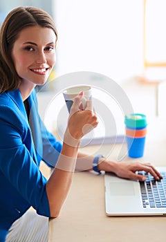 Attractive smiling business woman sitting at office desk, holding a cup of coffee