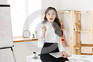 Attractive smiling business woman sitting at office desk, holding a cup of coffee, she is relaxing and happy