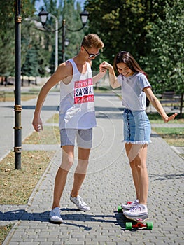 An attractive smiling fellow teaching a girl riding on a longboard in a park on a natural blurred background.