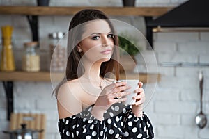 Attractive smiling girl in a polka dot dress drinks coffee and sitting on the wooden table in the cozy kitchen at the morning.