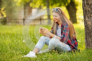 Attractive smiling girl typing on cell phone in summer city park. Modern happy woman with a smartphone, outdoor