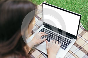 Attractive smiling girl works remotely behind a close-up laptop in a city park, lying on a plaid. A place for text. mock-up