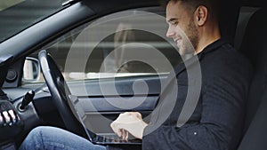 Attractive smiling man typing laptop computer while sitting inside his car outdoors