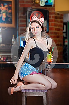Attractive smiling pinup woman in denim shorts sitting on bar stool and drinking lemonade